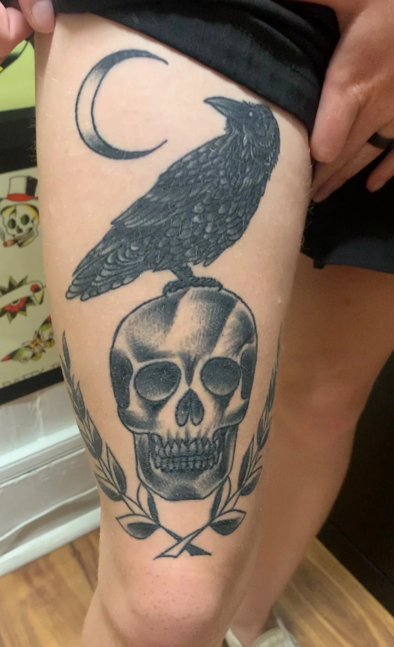 Black and grey Traditional thigh tattoo of a crow standing on a skull beneath a crescent moon, framed by a laurel wreath, by Tim Goodrich at his Waikiki tattoo shop, Hawaii.