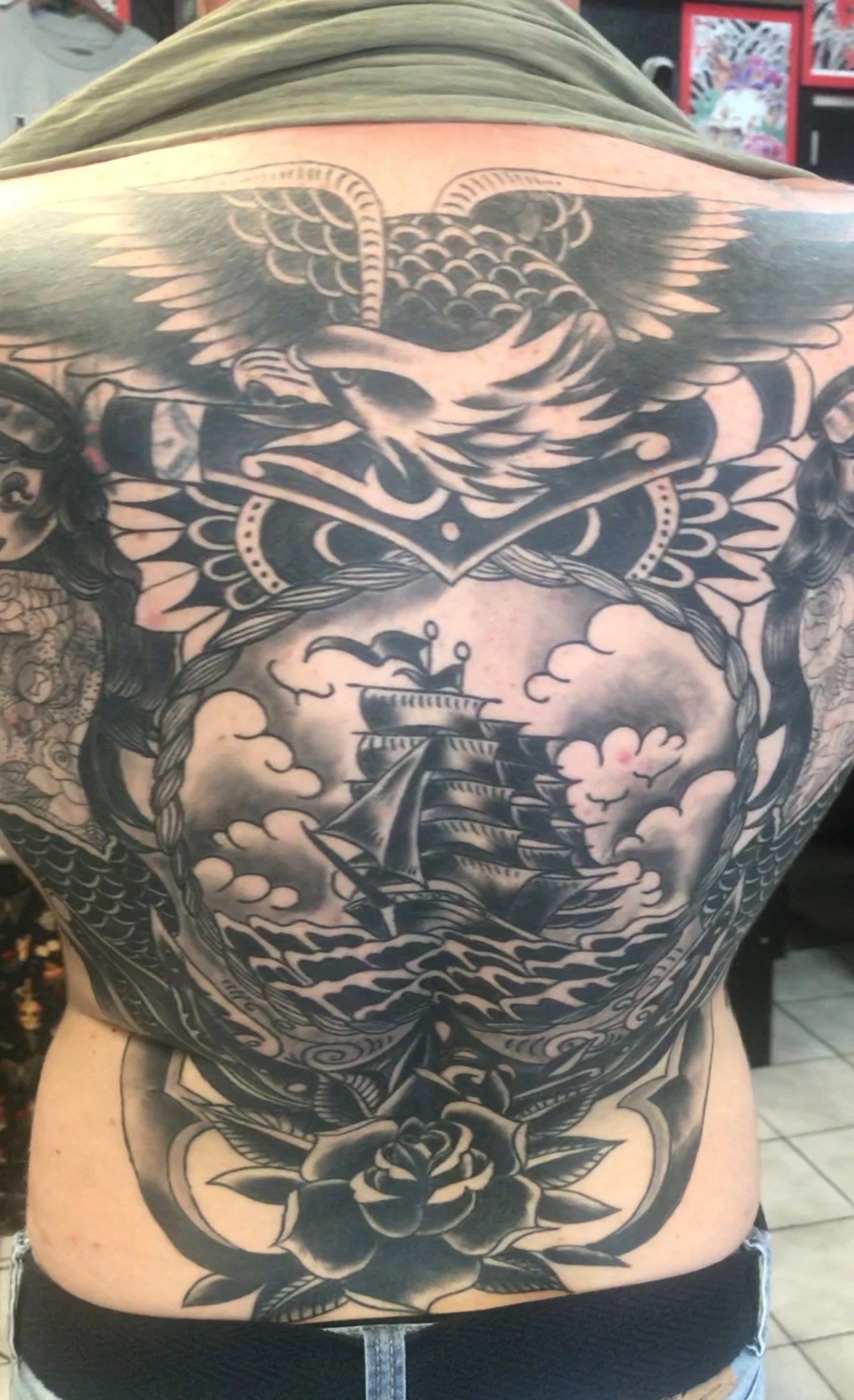 Black and grey Traditional full back tattoo: an eagle with spread wings at the top above an anchor, a clipper ship in an oval medallion in the center, and a rose at the low back, by Tim Goodrich at his Waikiki tattoo shop, Hawaii.