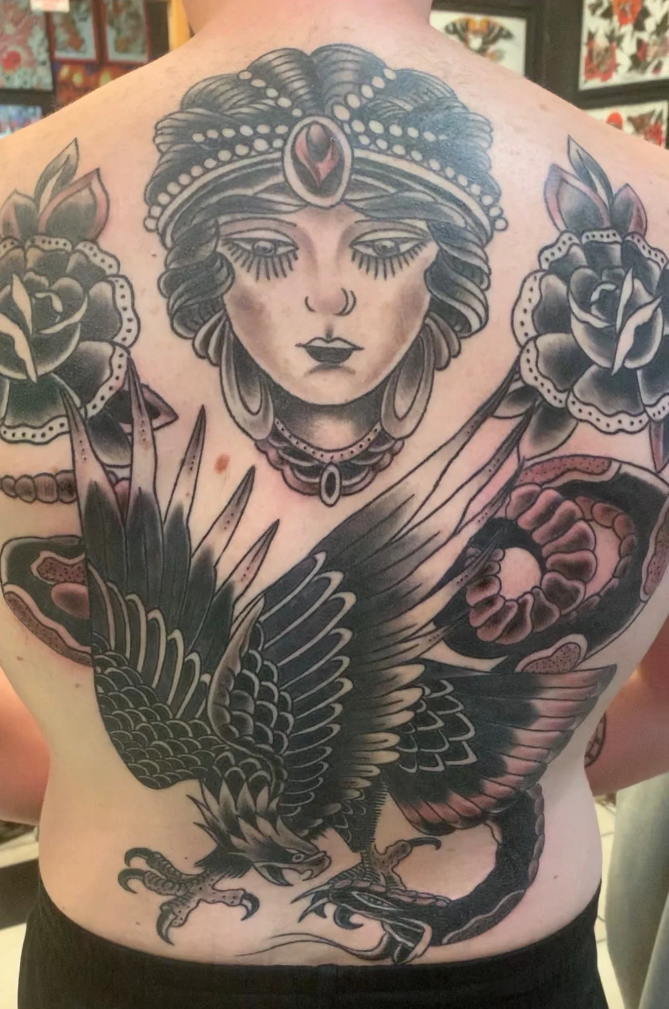 Black and grey Traditional full back tattoo of a gypsy queen in a crown with twin roses flanking, an eagle below and a snake coiling at the right side, by Tim Goodrich at his Waikiki tattoo shop, Hawaii.