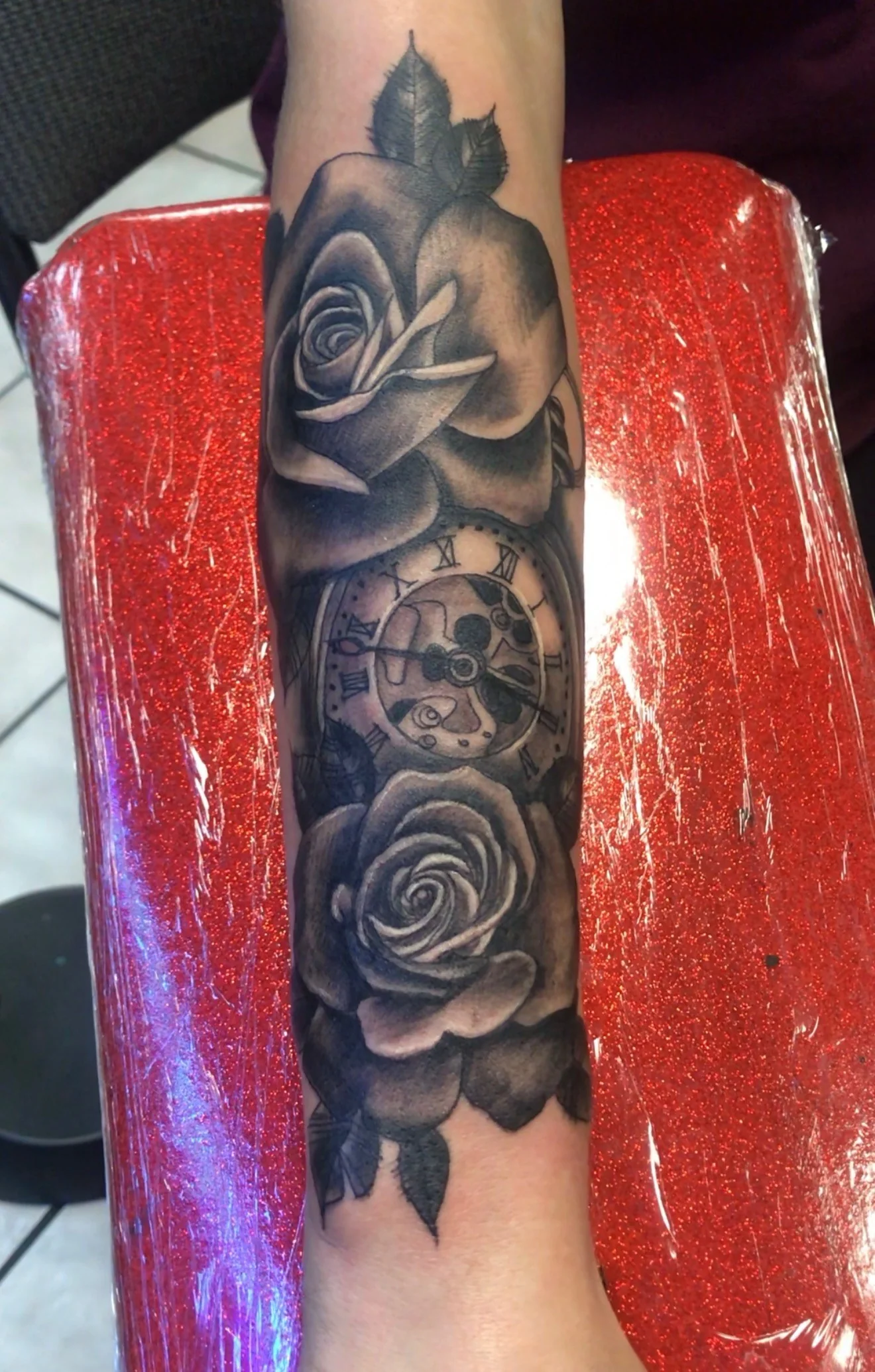 Black and grey tattoo of two open roses bracketing a Roman-numeral pocket watch, on the forearm, by Tim Goodrich at his Waikiki tattoo shop, Hawaii.