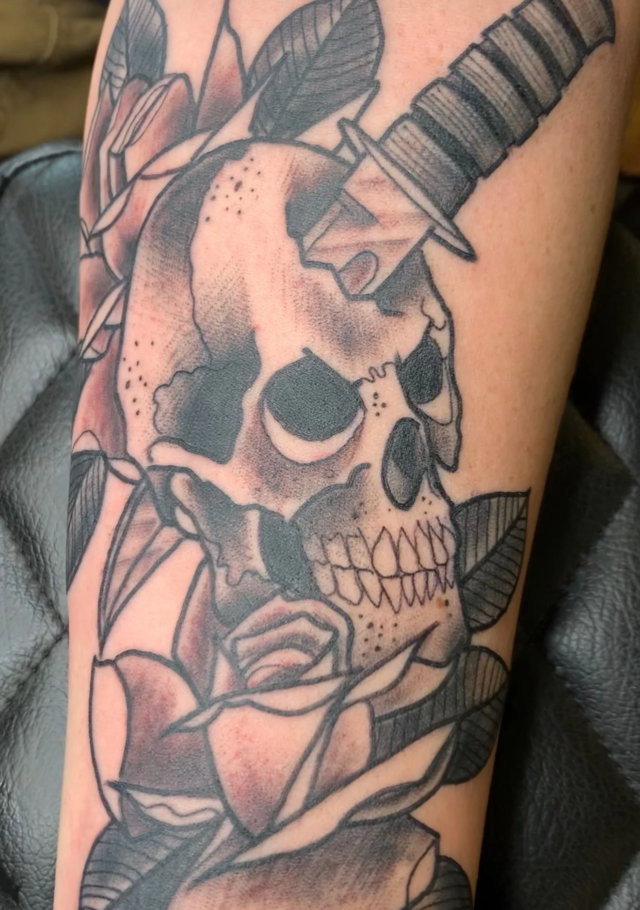 Black and grey Traditional forearm tattoo of a skull pierced through the top by a dagger, with a rose at the base and feathered leaves around, by Tim Goodrich at his Waikiki tattoo shop, Hawaii.