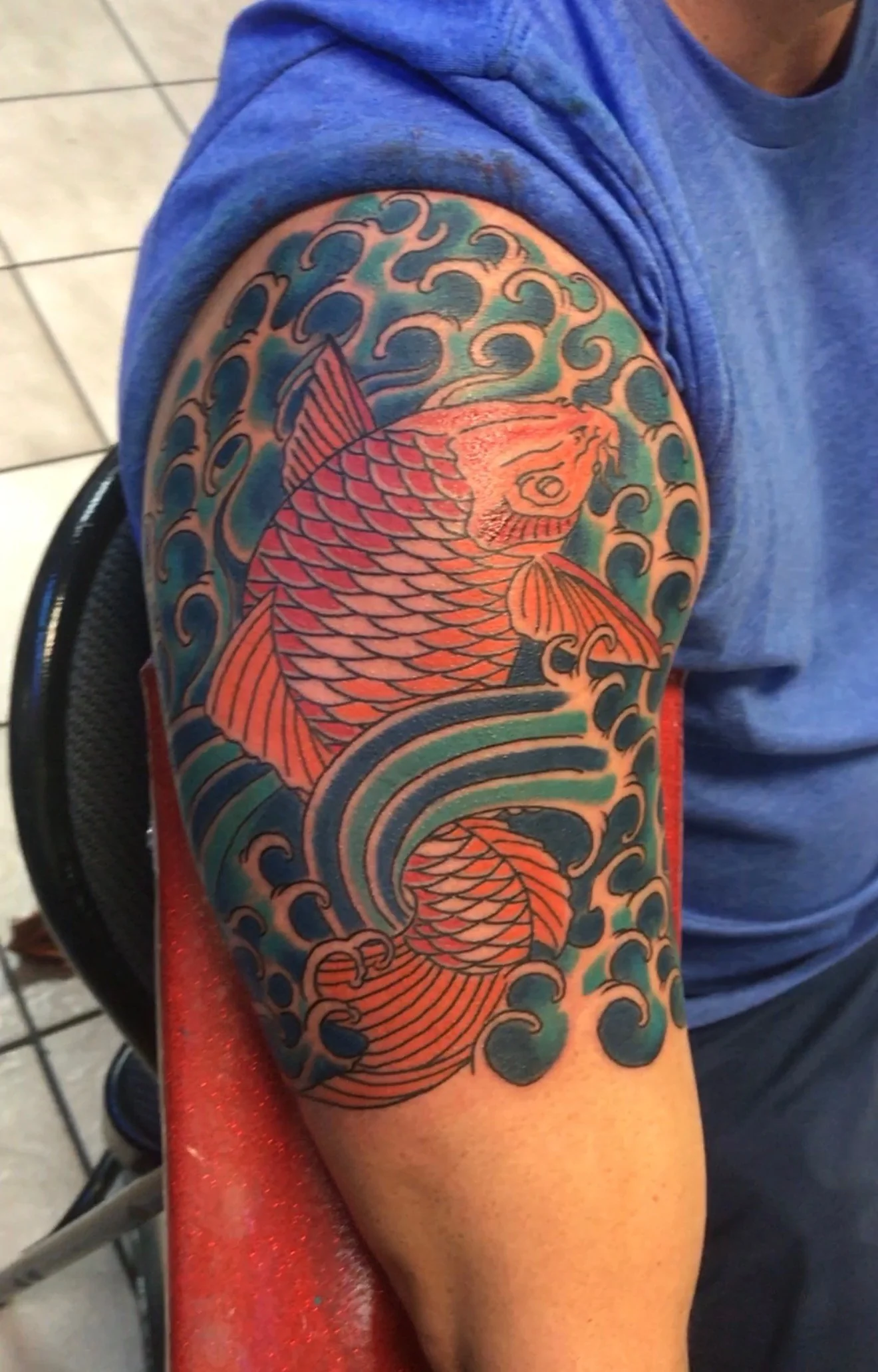 Japanese Wabori half-sleeve tattoo of a saturated red koi climbing through teal-and-black wabori waves on the upper arm, by Tim Goodrich at his Waikiki tattoo shop, Hawaii.
