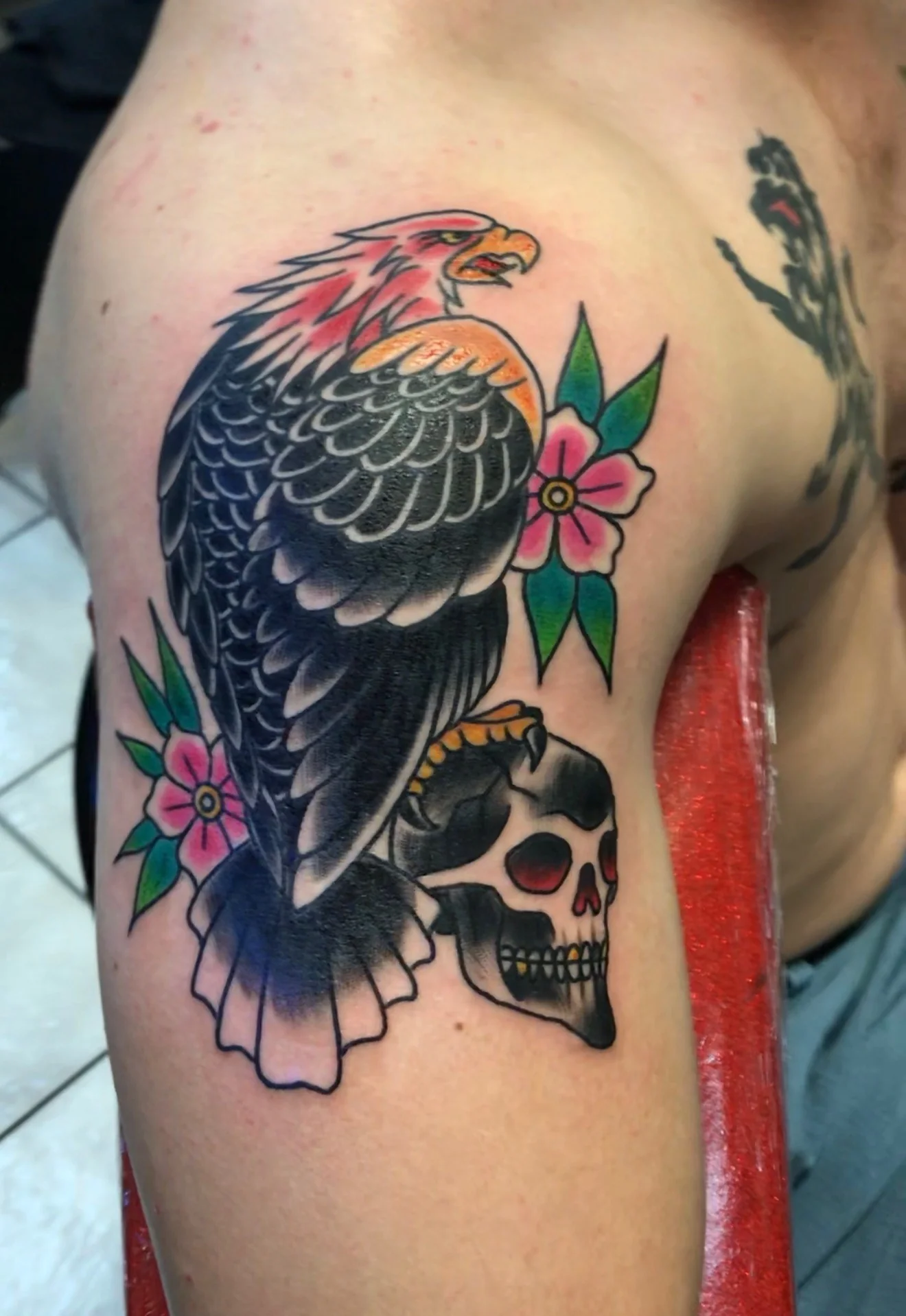 American Traditional color tattoo of an eagle perched on a skull flanked by pink Sailor Jerry flowers, on the shoulder, by Tim Goodrich at his Waikiki tattoo shop, Hawaii.